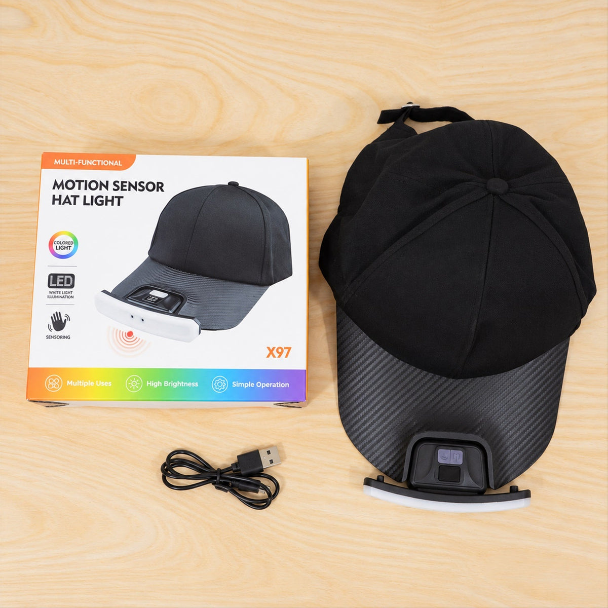 A black baseball cap with an LED light, its product box, and a coiled USB charging cable laid out on a light wooden surface.