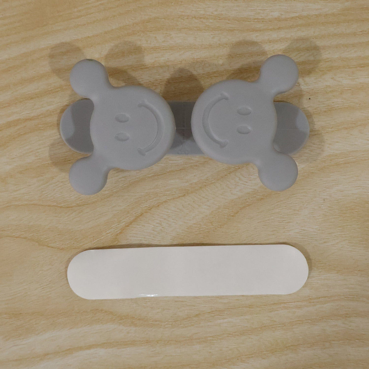 A gray silicone cord organizer with two smiling faces and a white adhesive strip on a light wood surface.