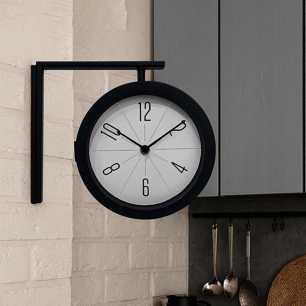 A black double-sided wall clock with a white face and black numbers is mounted on a white brick wall in a kitchen, with dark