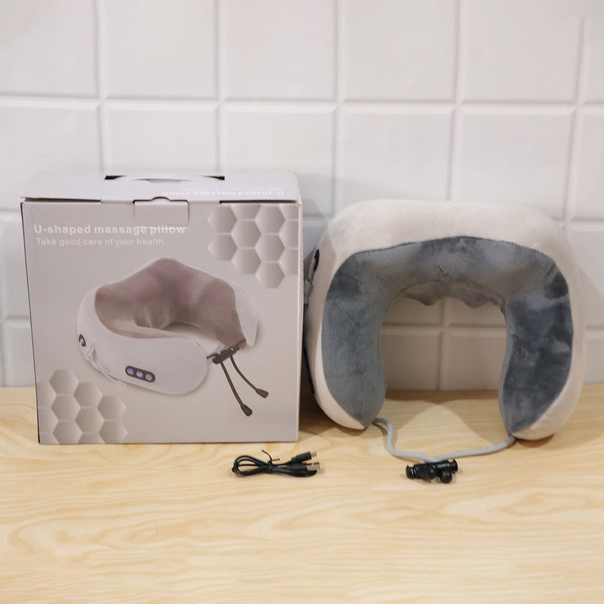 A grey and white U-shaped neck massage pillow, its packaging box, and a charging cable on a wooden surface.