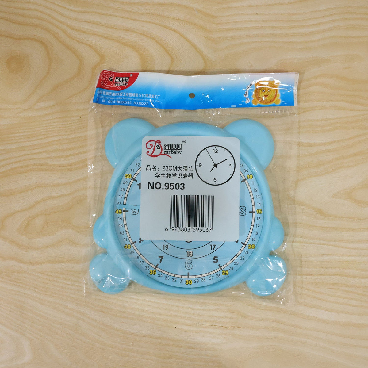 A light blue bear-shaped learning clock is sealed in clear plastic packaging with a white label on a wooden surface.
