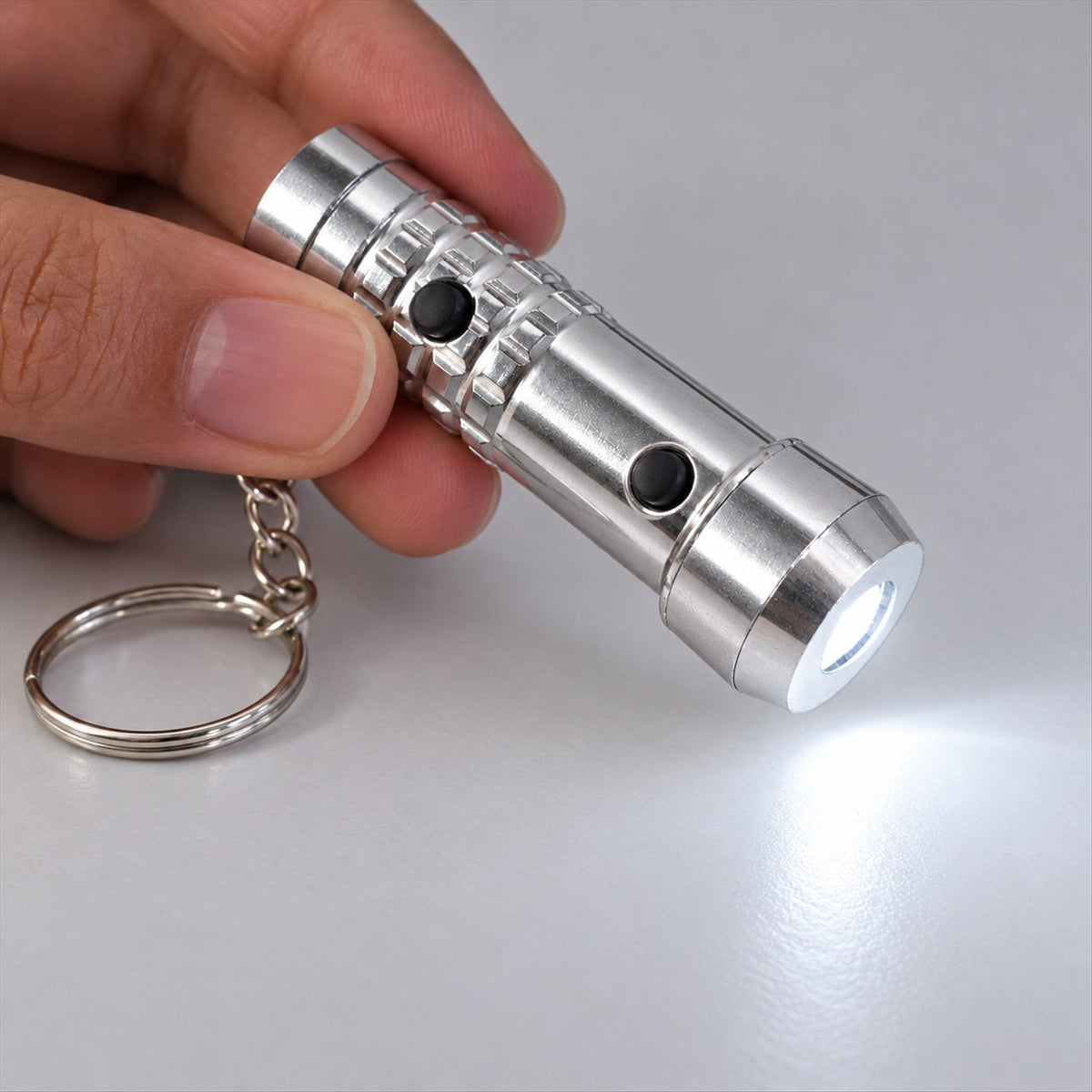 A hand holds a silver keychain flashlight with two black buttons, emitting a bright white light onto a light gray surface.