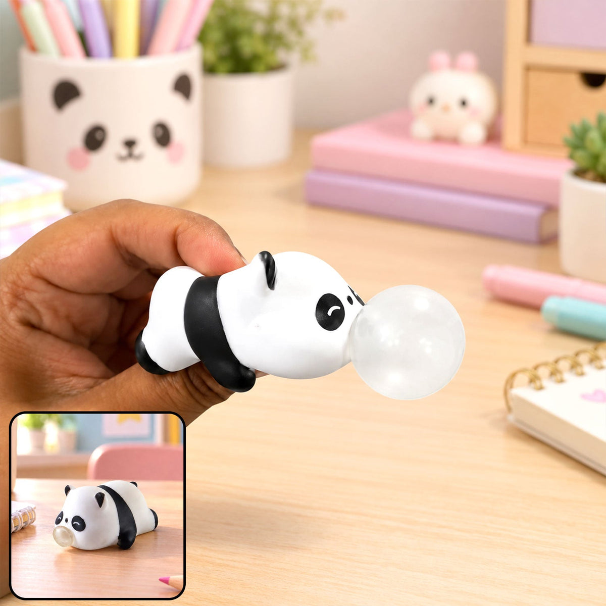 A hand holds a white and black panda squishy toy with a clear bubble emerging from its mouth, on a wooden desk.