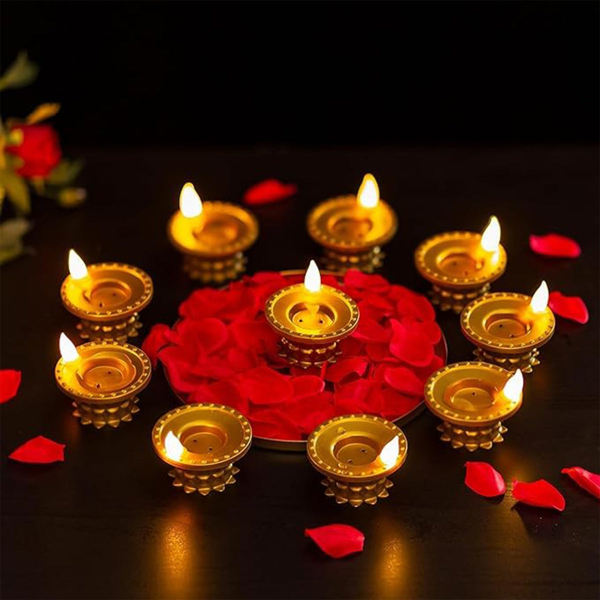 Nine gold plastic LED diyas with glowing flames arranged around a bed of red rose petals on a dark surface.