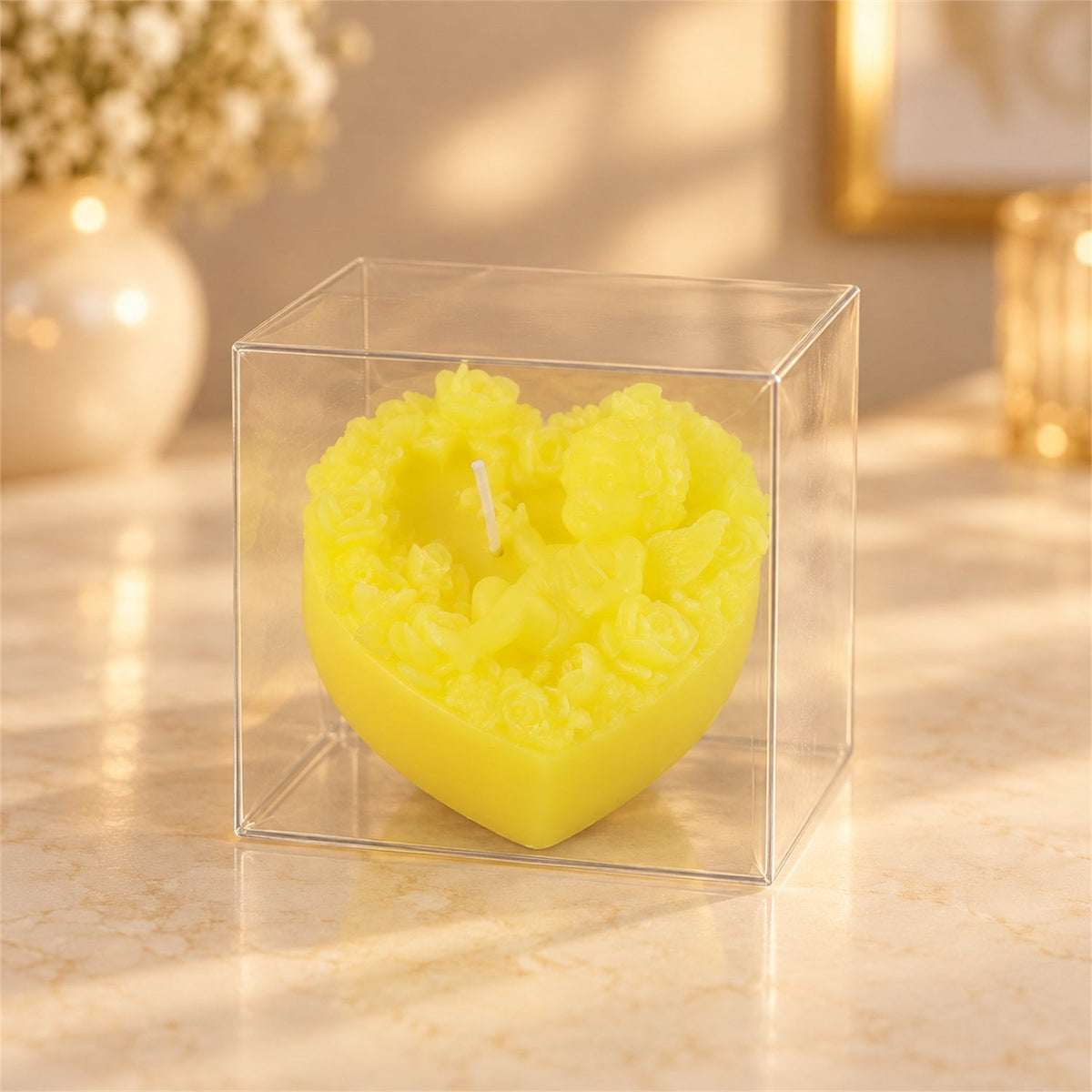A vibrant yellow heart-shaped candle with intricate floral and cherub details, encased in a clear plastic box.