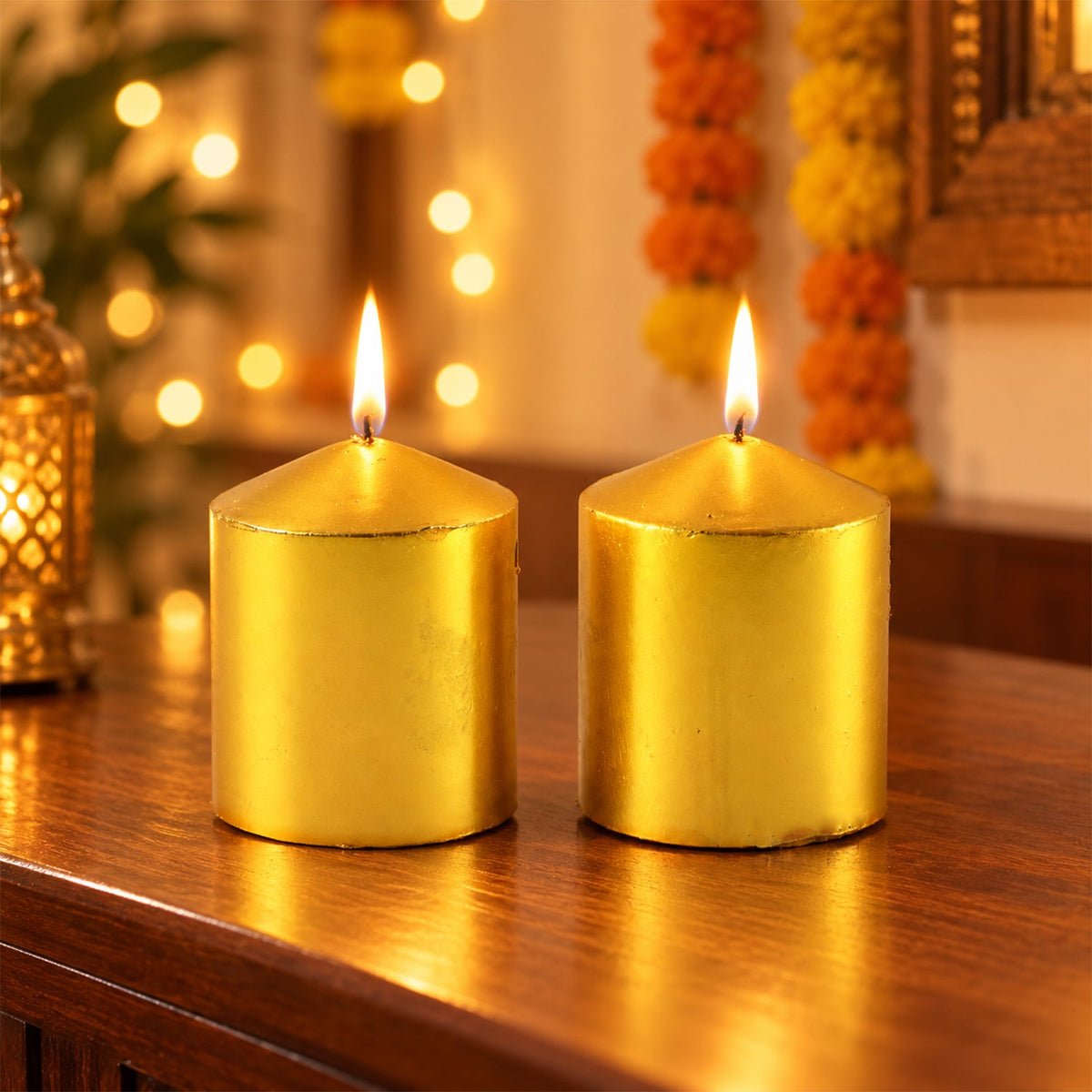 Two lit golden metallic pillar candles on a wooden table, with blurred warm lights in the background.
