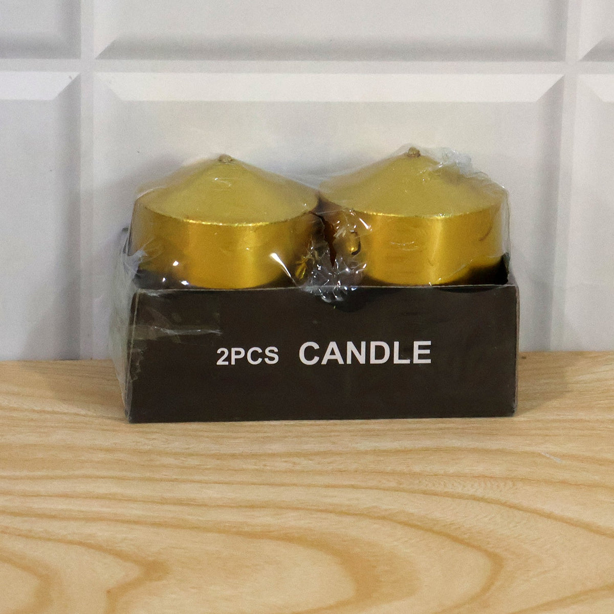 Two golden metallic pillar candles in clear plastic packaging within a black box labeled "2PCS CANDLE" on a wooden surface.