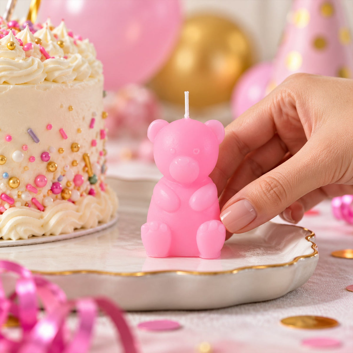 A hand places a pink teddy bear candle on a plate next to a small birthday cake with sprinkles.