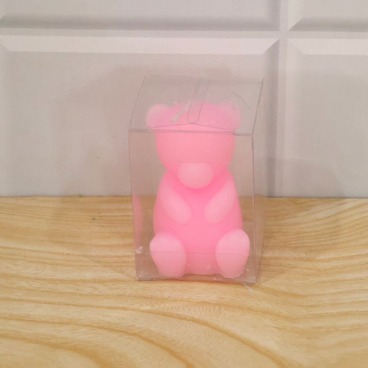 A pink teddy bear candle is packaged inside a clear plastic box on a light wooden surface.
