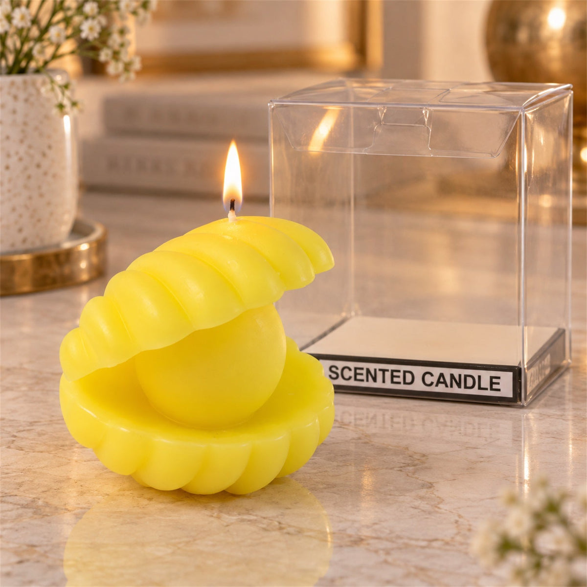 A lit yellow shell-shaped candle with a pearl center sits beside its clear plastic packaging on a marble surface.