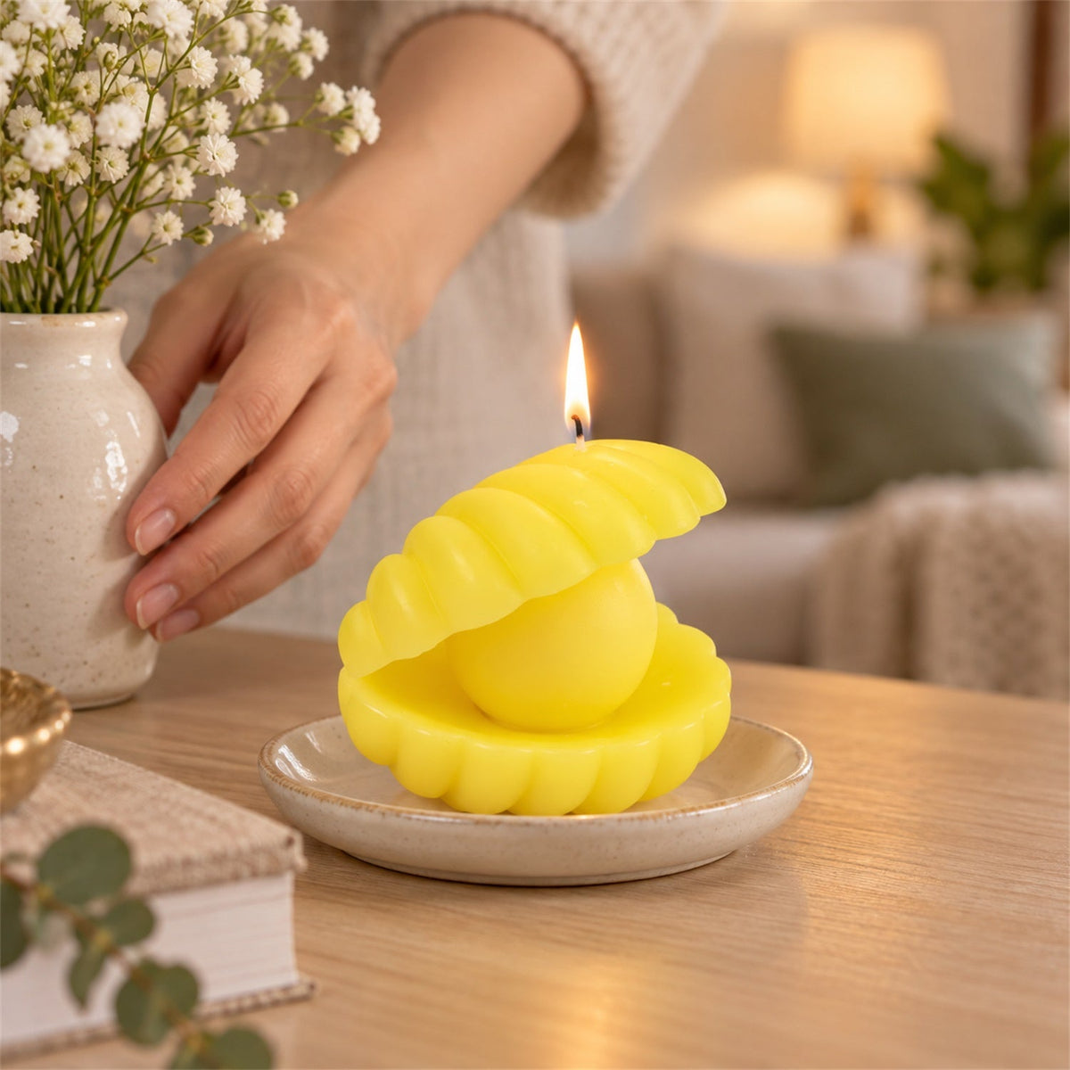 A person's hands adjust a vase of white flowers next to a lit yellow shell candle on a small ceramic plate.