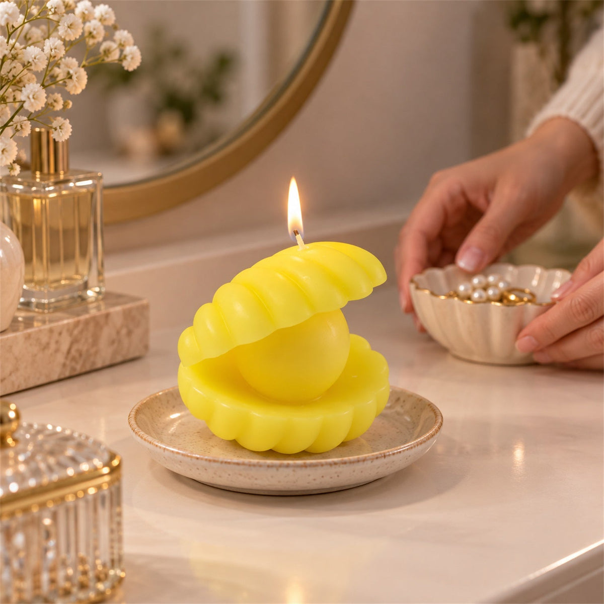 A yellow shell candle on a small plate sits on a vanity, while hands reach for a decorative bowl of pearls.