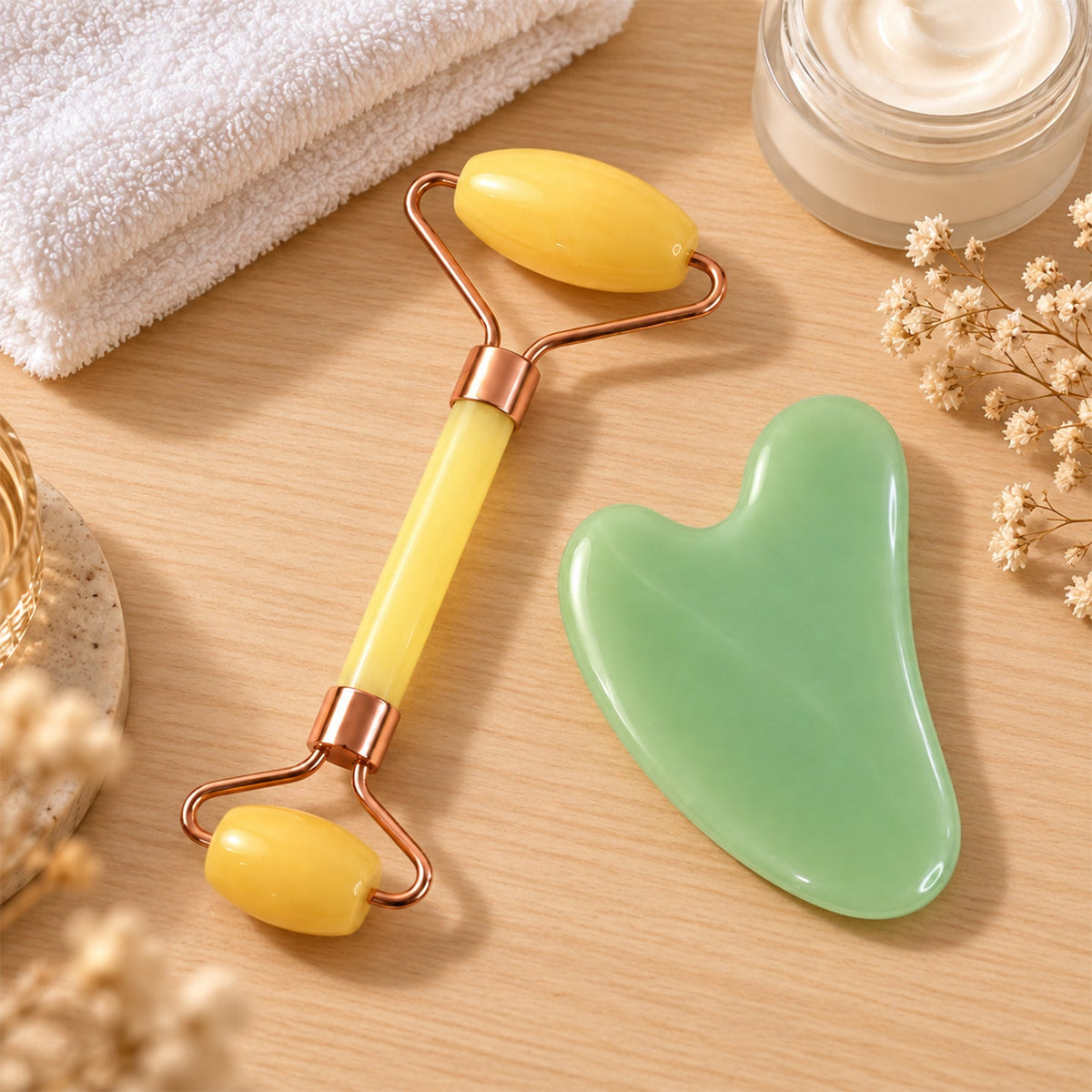 A yellow facial roller and green gua sha tool rest on a wooden surface next to a towel and cream jar.