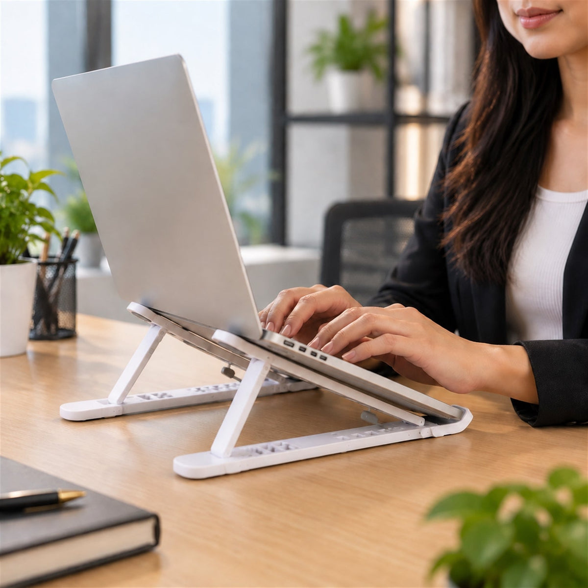 Woman typing on a silver laptop elevated by a white stand on a wooden office desk.