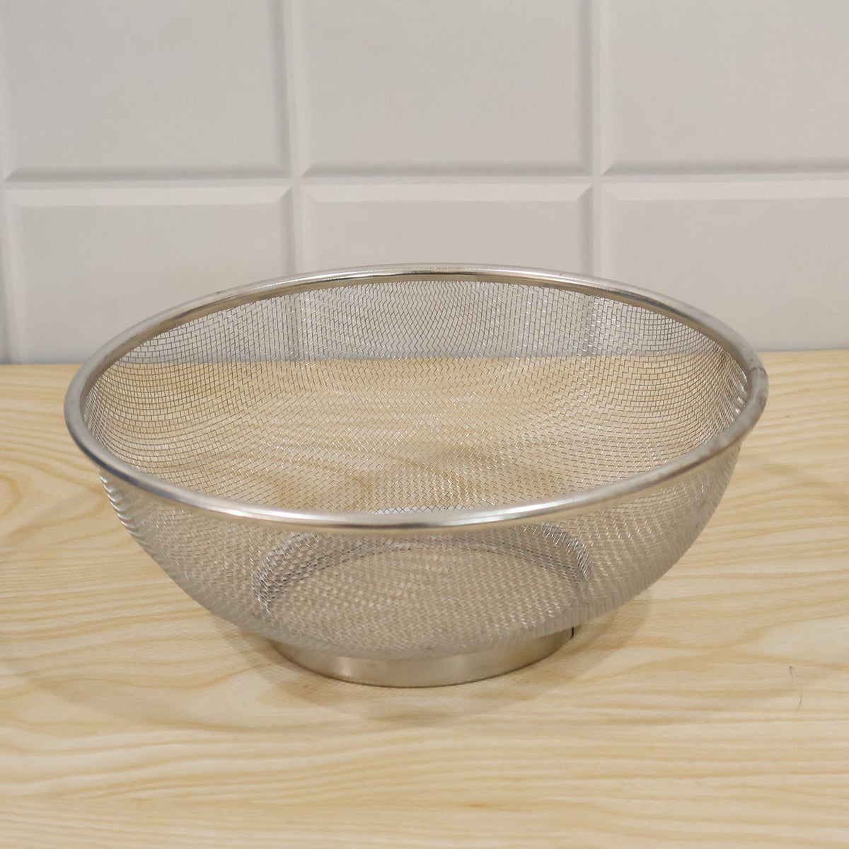 A silver stainless steel mesh strainer bowl with a solid base sits on a light wooden surface.