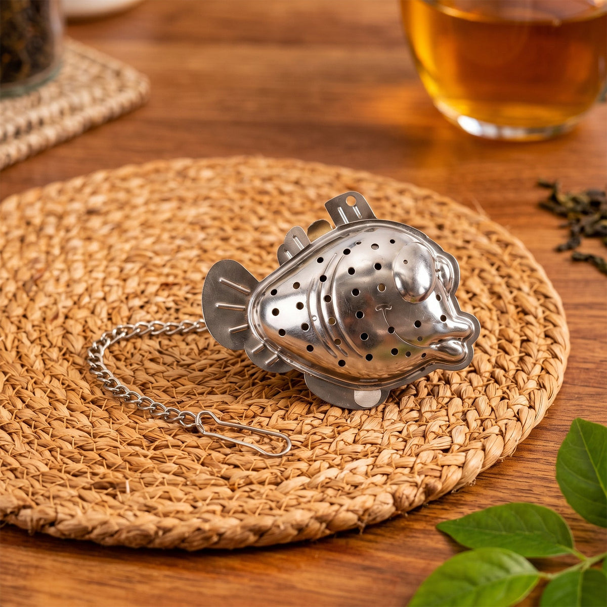 A metallic fish-shaped tea infuser with a chain and hook rests on a round woven placemat on a wooden surface.