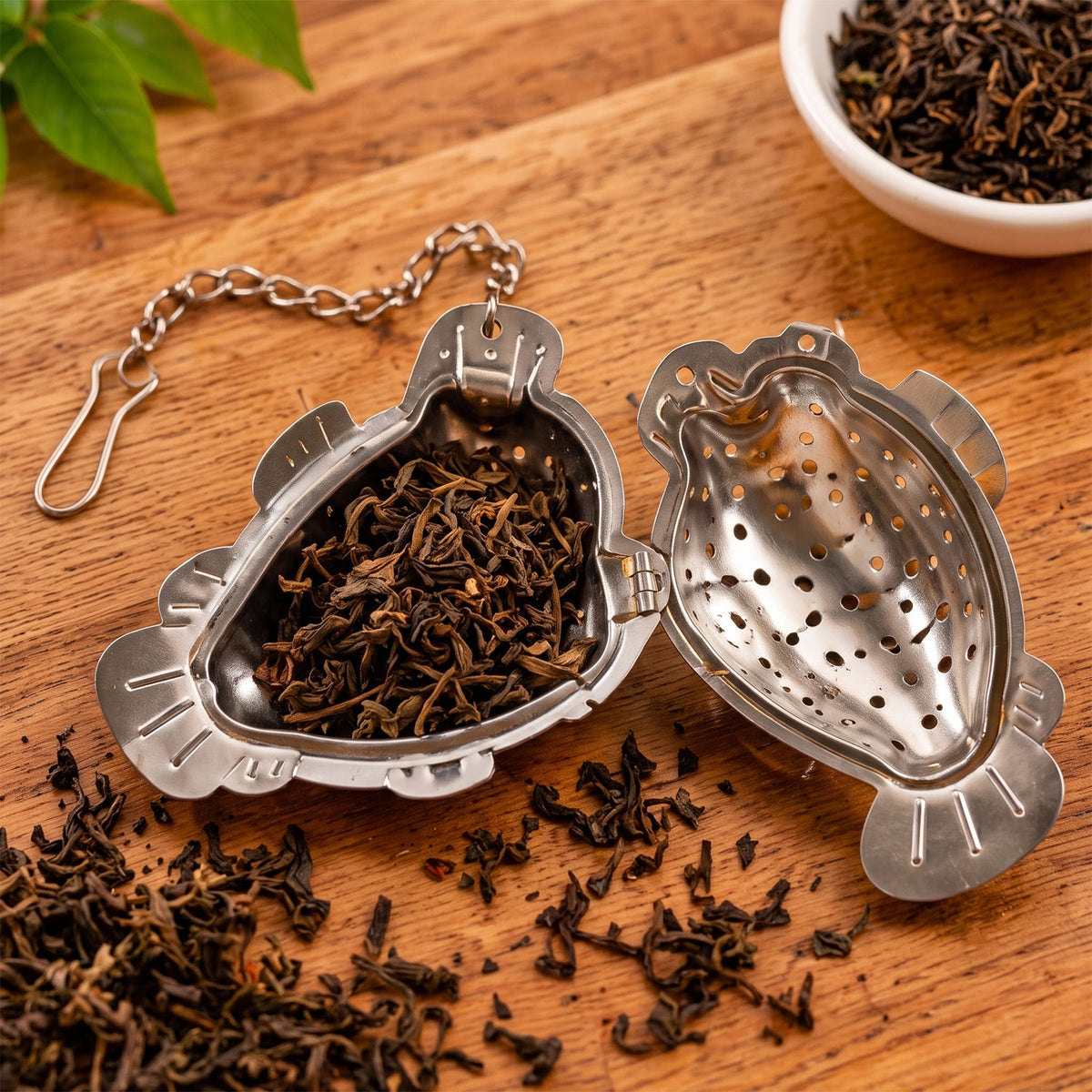 An open silver fish-shaped tea infuser filled with loose dark tea leaves, next to more scattered leaves on a wooden table.