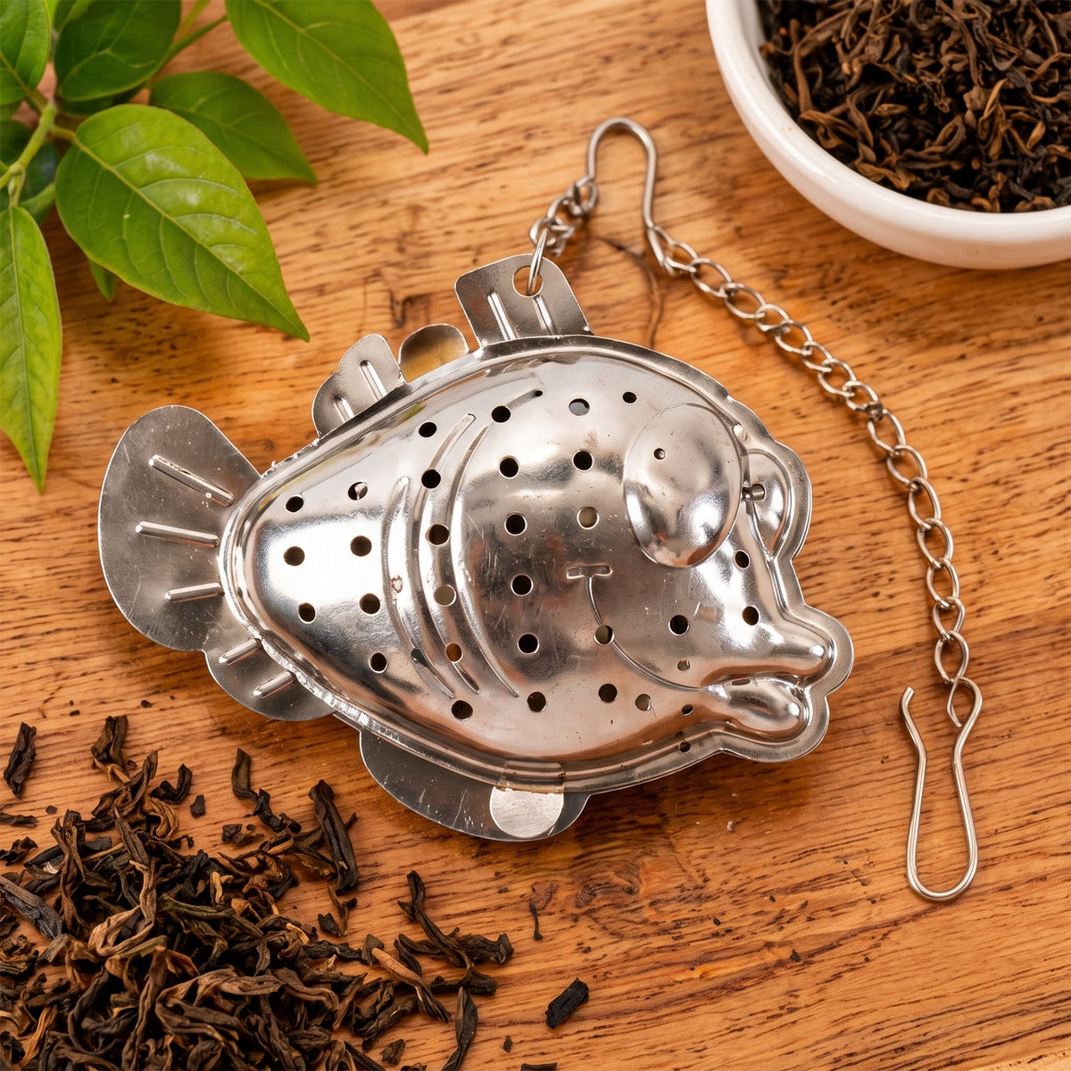A closed silver fish-shaped tea infuser with its chain and hook, surrounded by loose dark tea leaves on a wooden surface.
