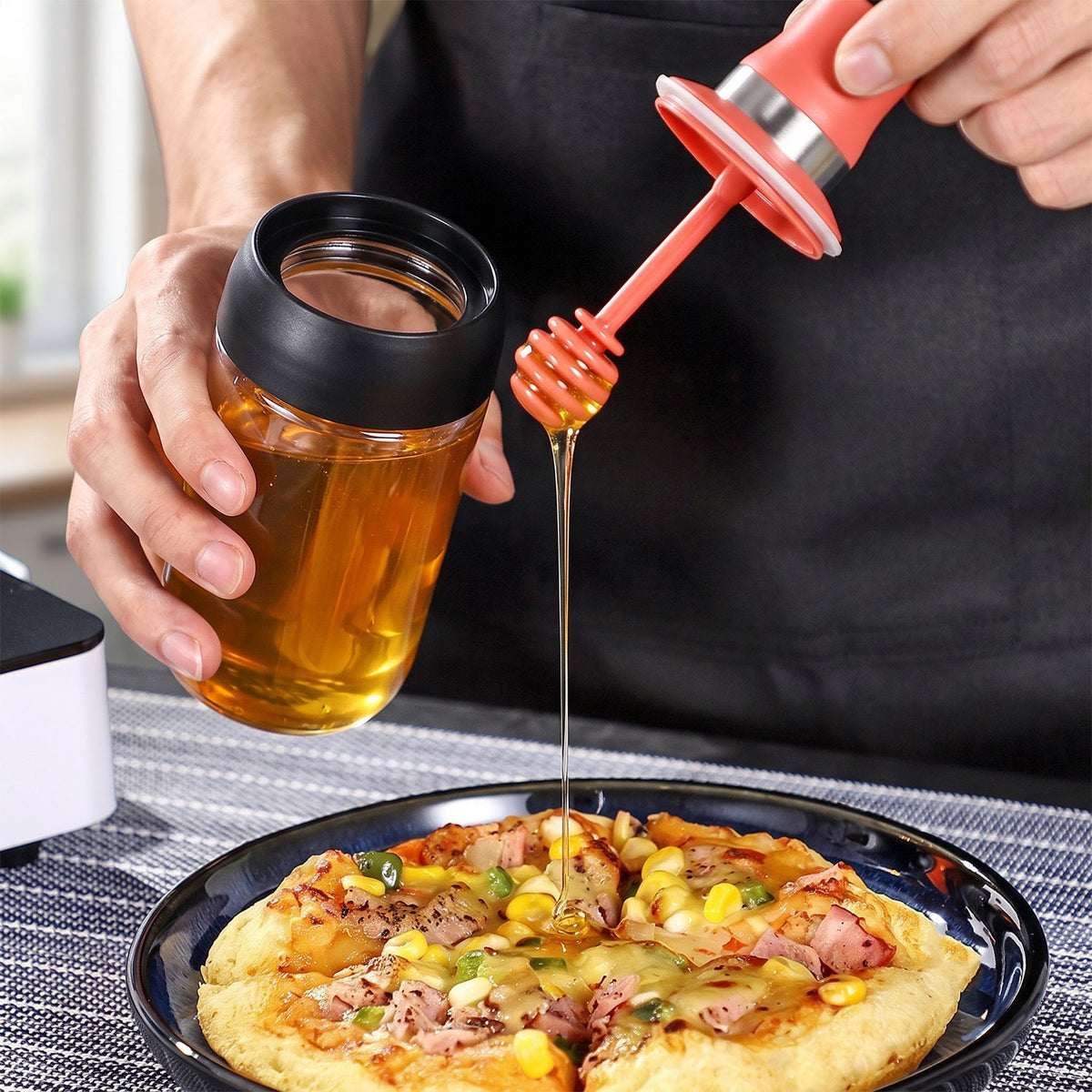 A person drizzles honey from a glass dispenser with a red silicone dipper onto a pizza slice. â€“ view 8