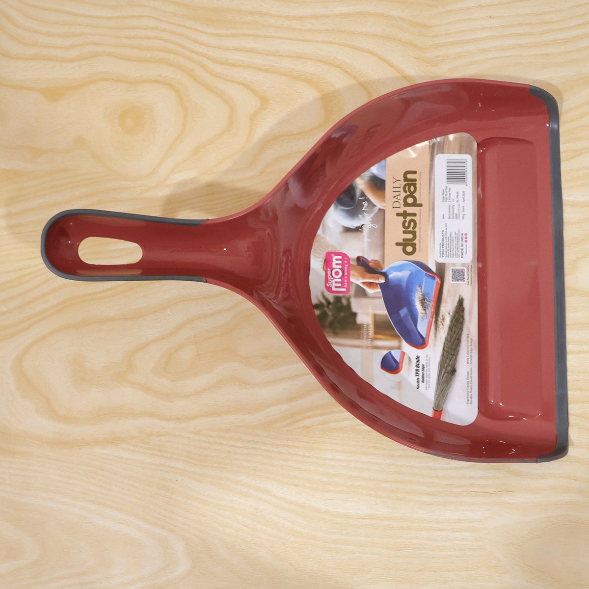 A red and grey dustpan with a product label affixed to its surface lies on a light wooden background.