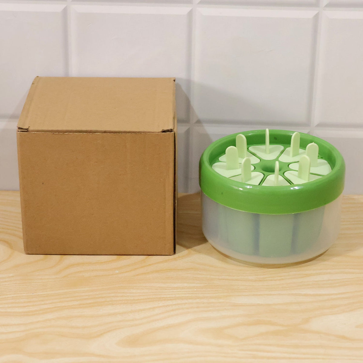A green and white round plastic ice cream mold with its eight stick lids, next to a plain brown cardboard box on a wooden