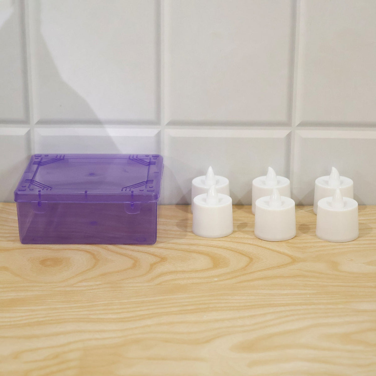 A purple translucent plastic box sits next to six white flameless tealight candles on a light wooden surface against a tiled
