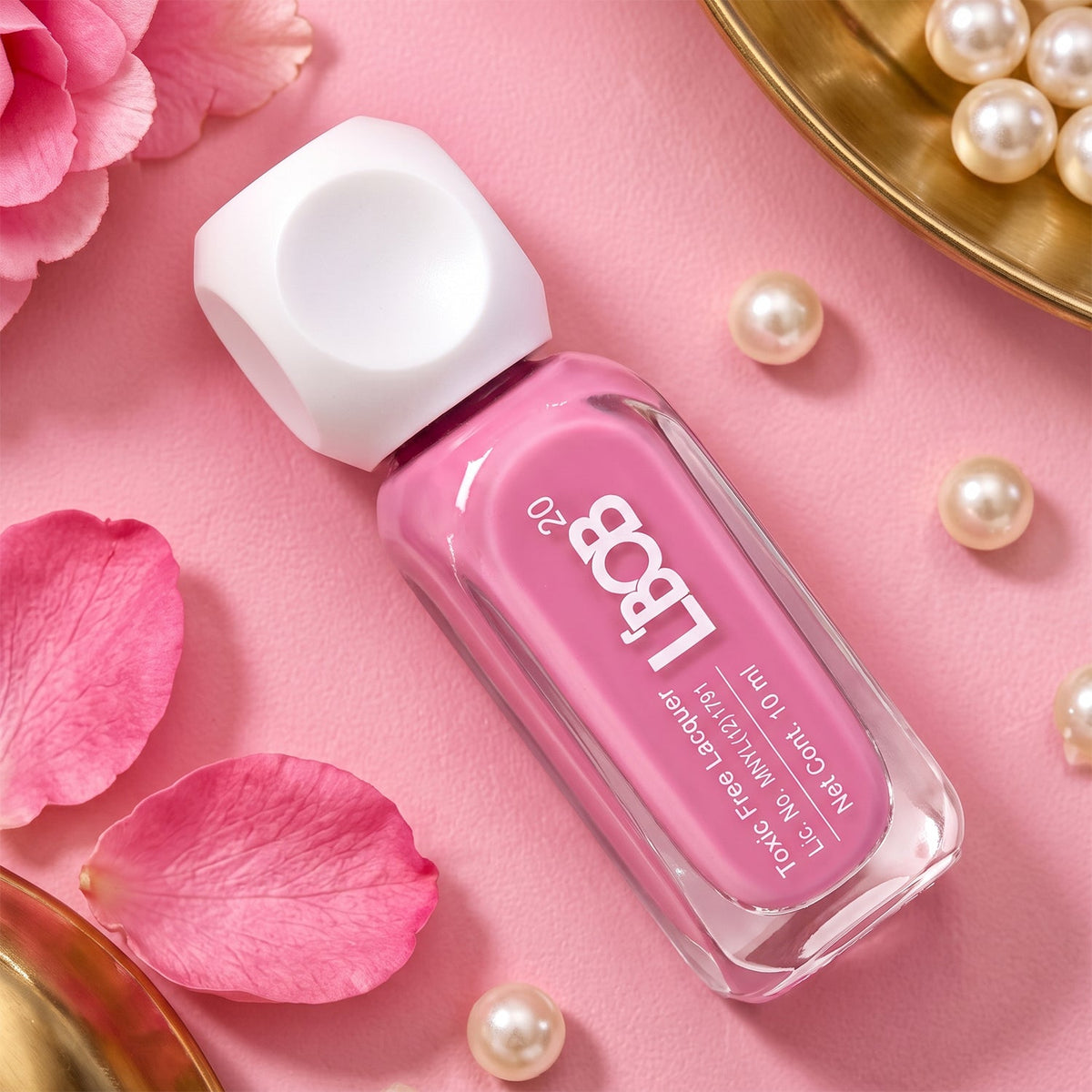 A pink nail polish bottle with a white cap lies diagonally on a pink surface, adorned with scattered pearls and petals.