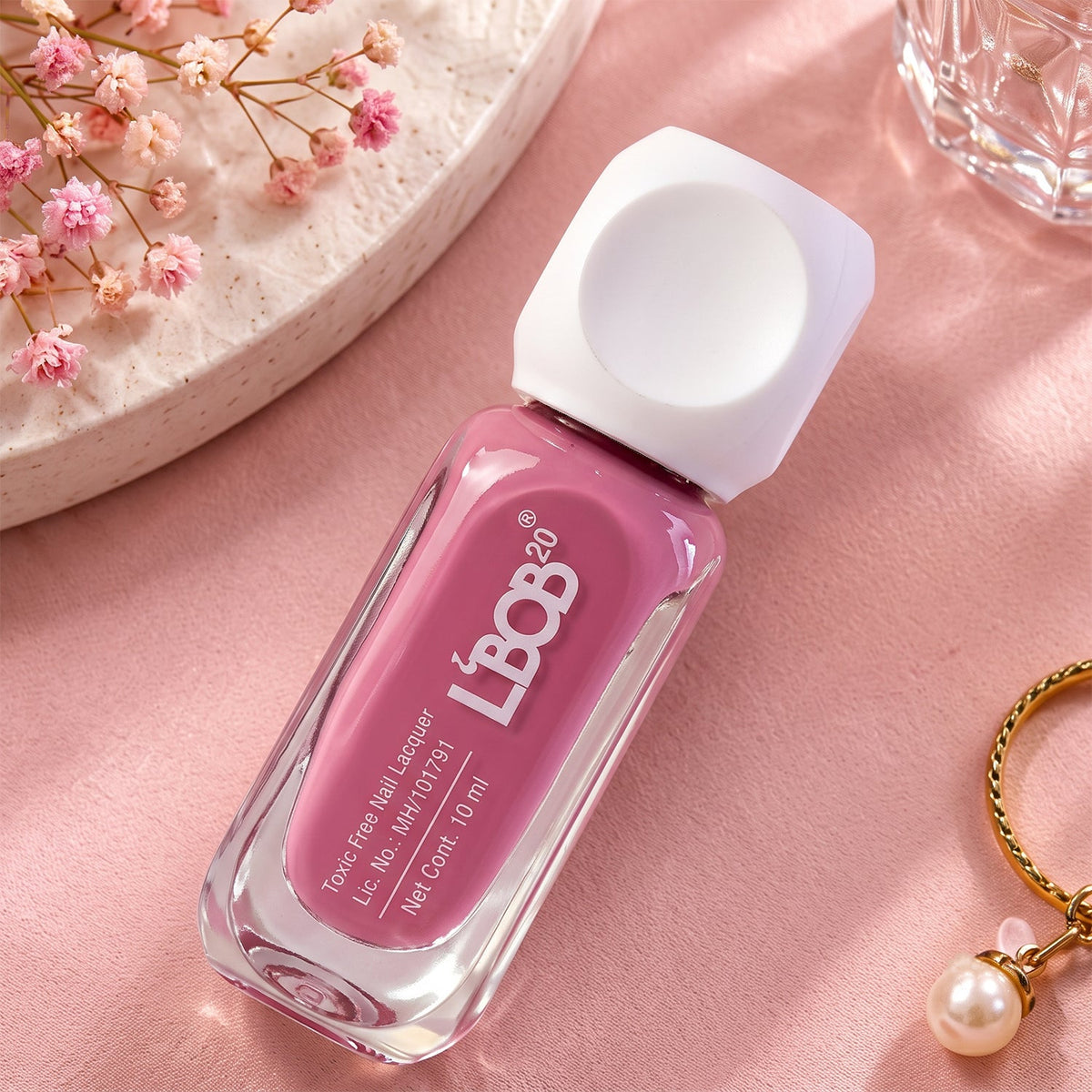 A mauve nail polish bottle rests on a pink surface next to a pearl charm and small flowers.