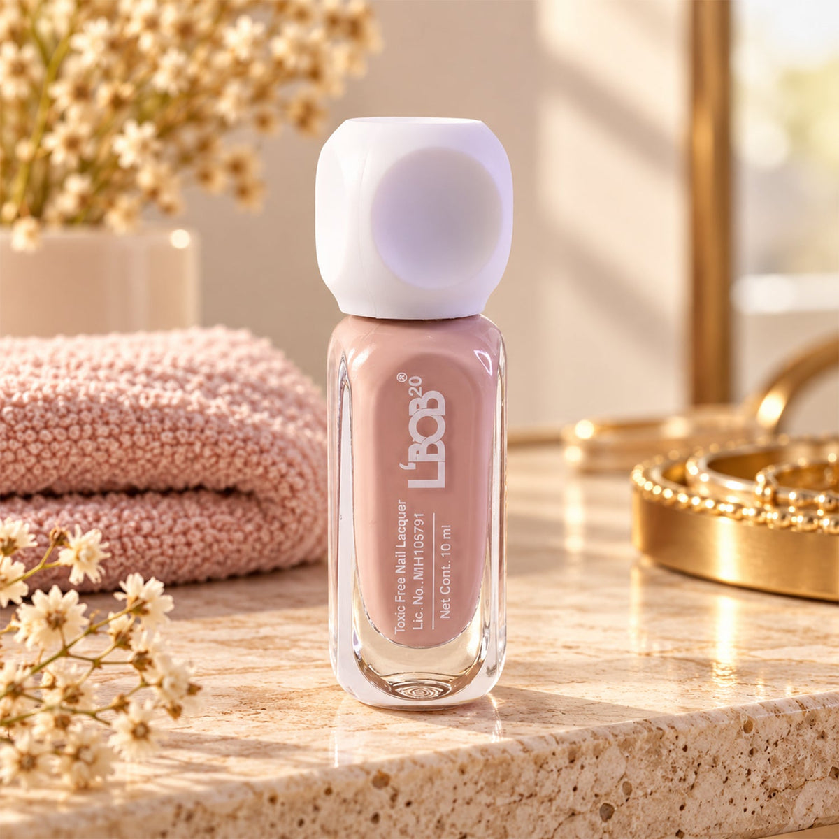 A bottle of nude pink nail polish on a marble surface with a folded pink towel and flowers in the background.