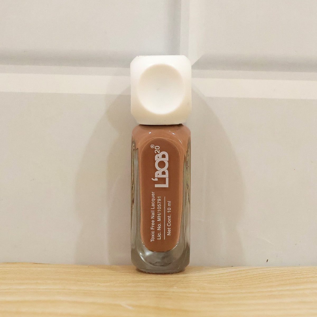 A small bottle of nude brown nail polish with a white cap stands on a wooden surface against a white door.