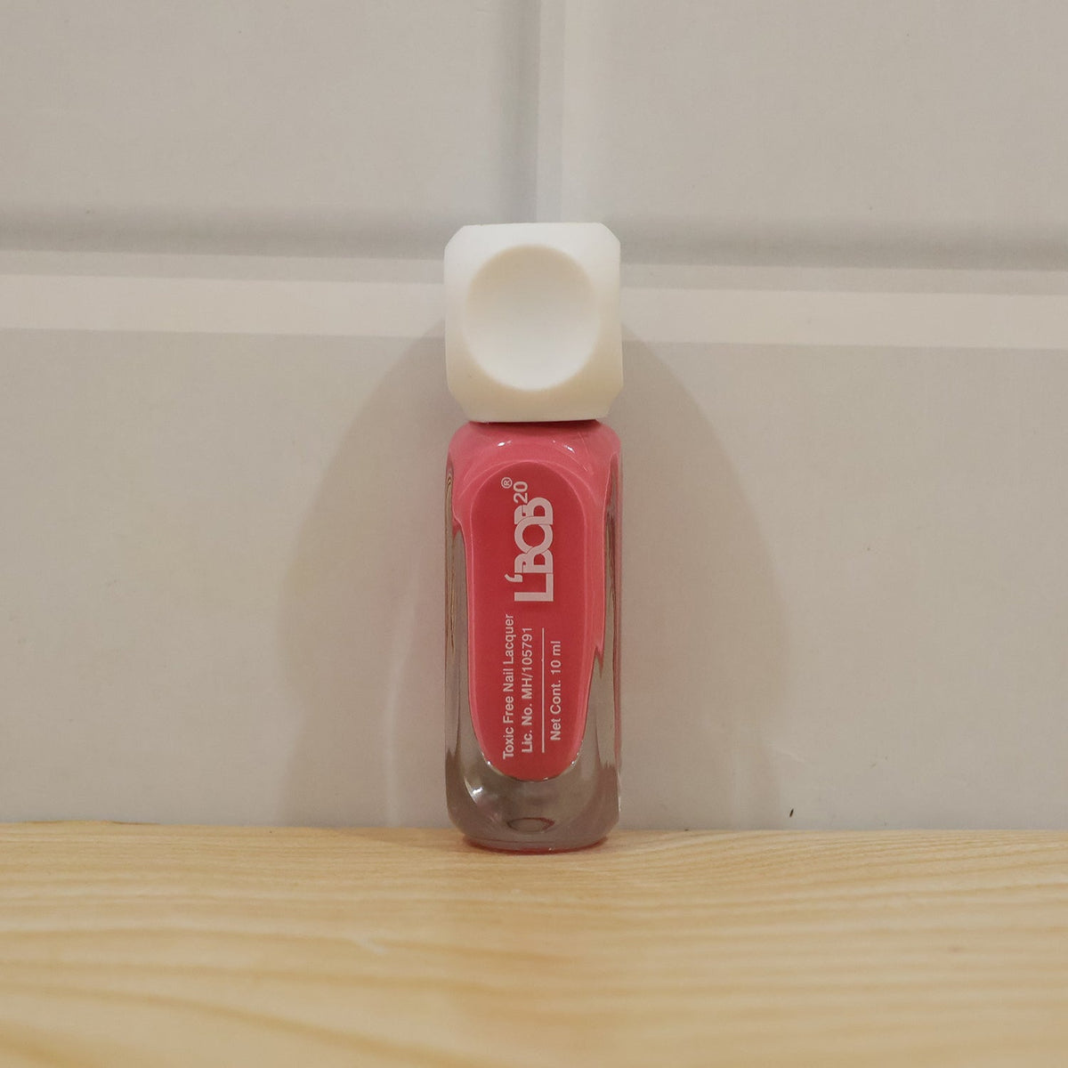 A pink nail polish bottle with a white cap stands upright on a light wooden surface against a white paneled wall.