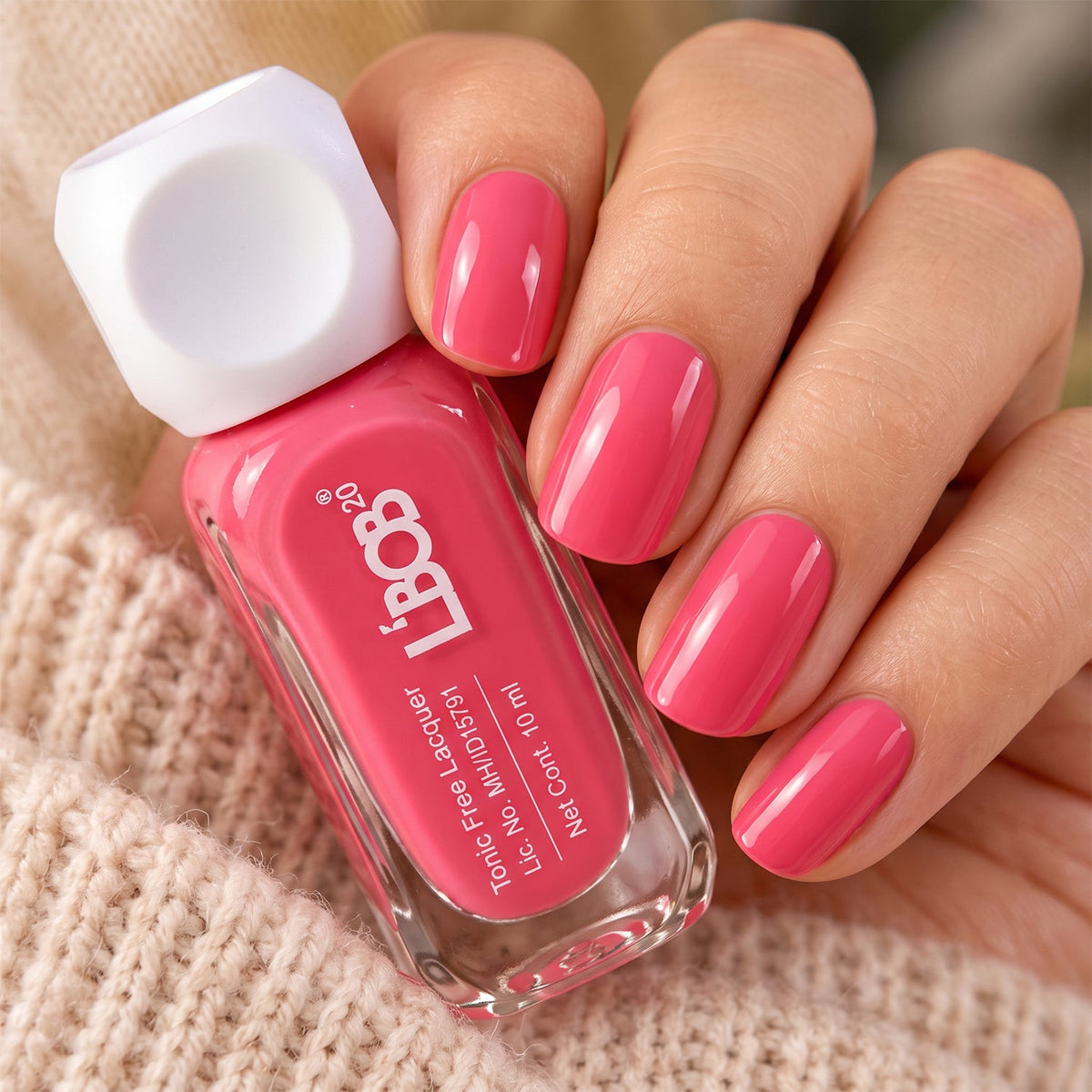 A hand with perfectly painted pink nails holds a bottle of matching nail polish, resting on a soft, knitted fabric.