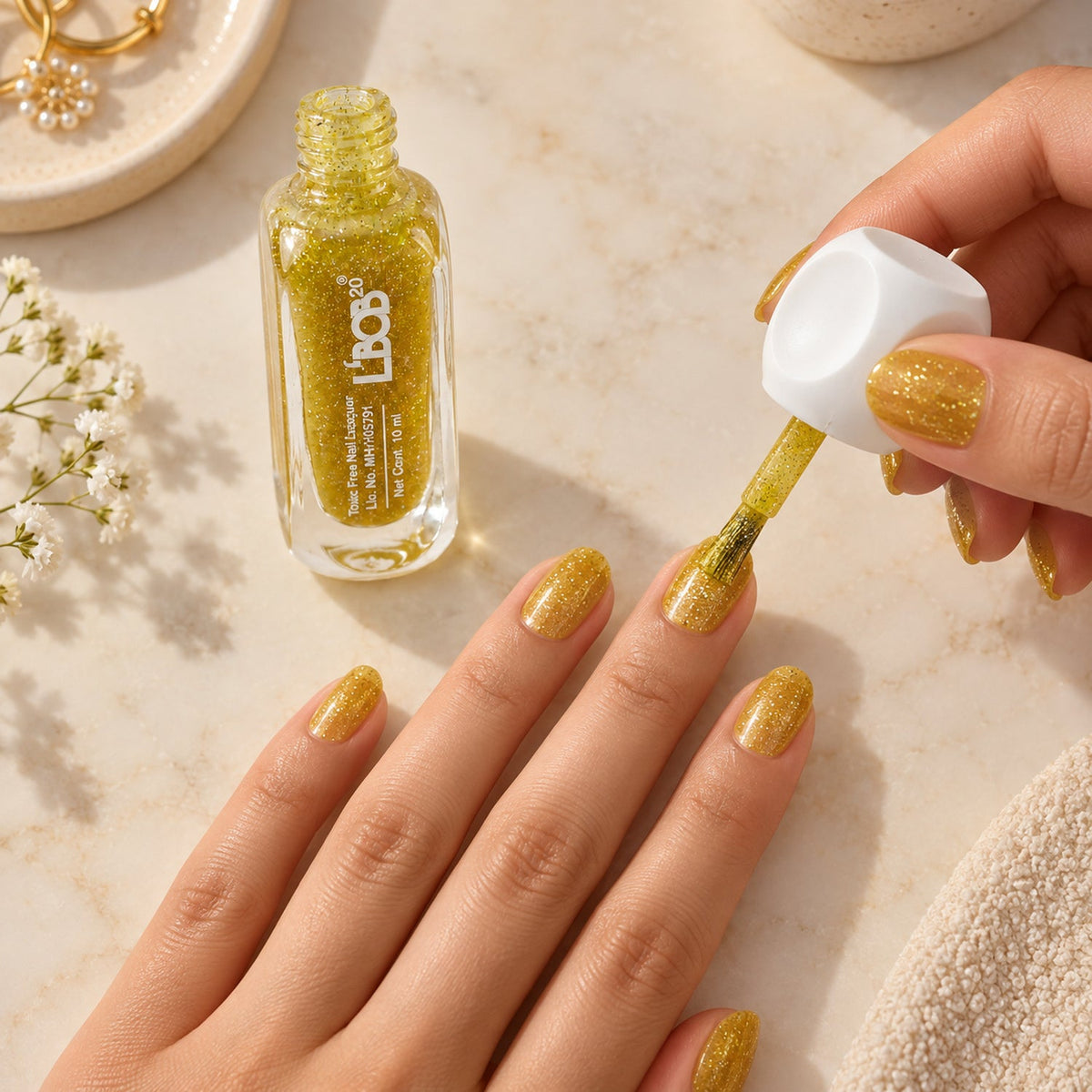A person applies gold glitter nail polish to their fingernails, with a bottle of polish and a white towel nearby.