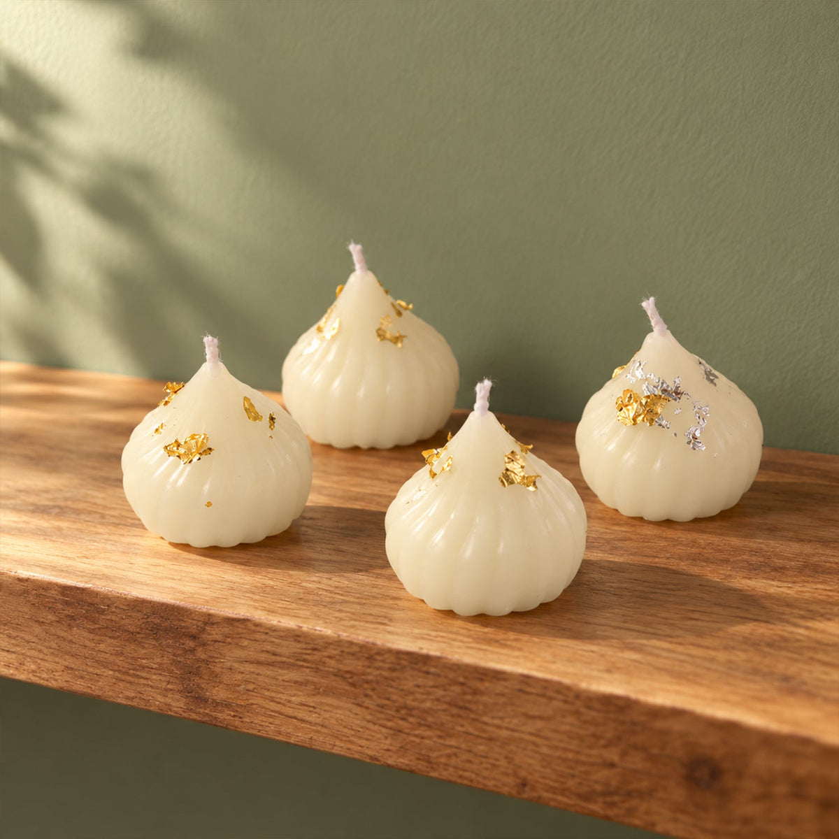 Four unlit modak-shaped white wax candles with gold and silver flakes on a light wooden shelf against a green wall.