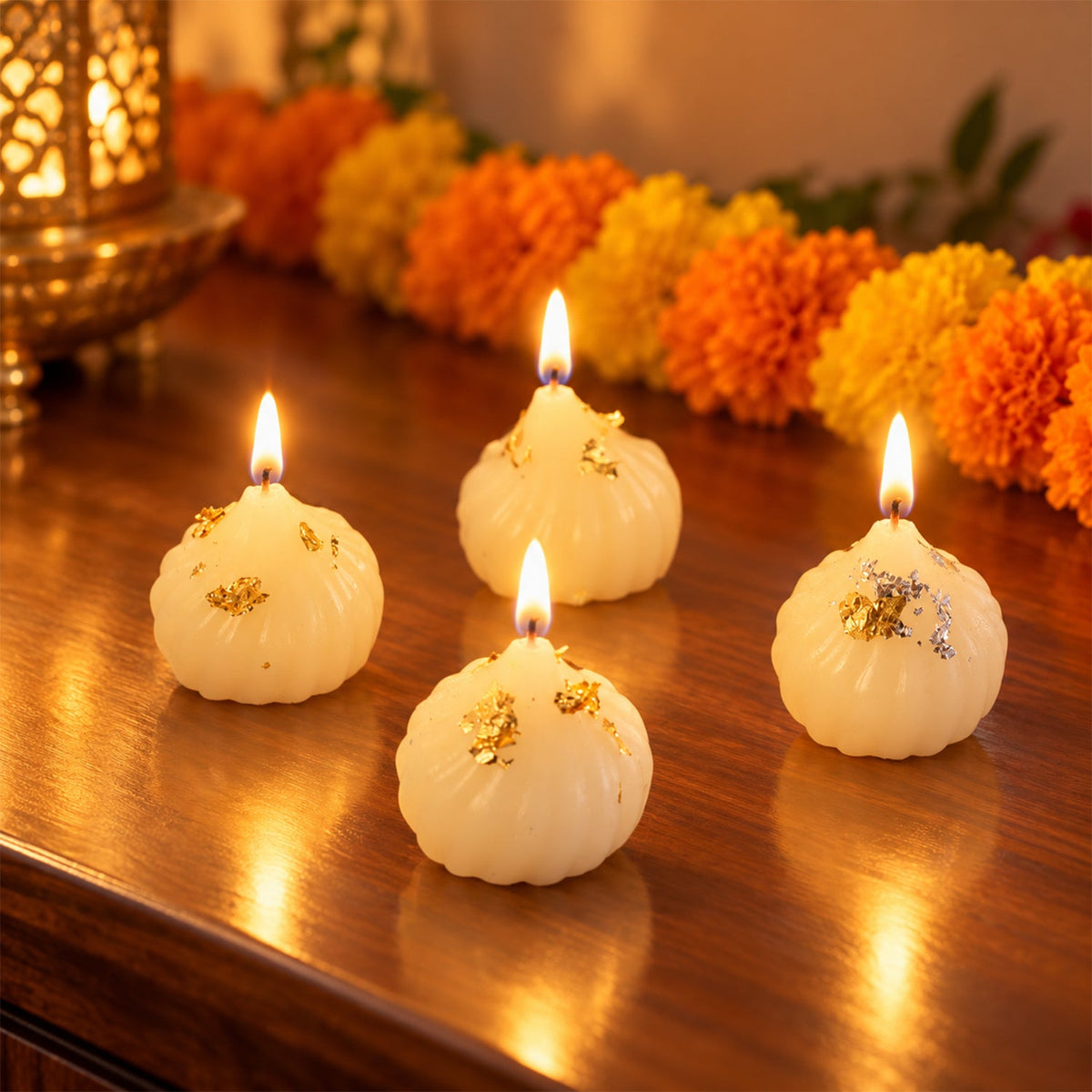 Four lit white modak-shaped candles with gold and silver flakes on a dark wooden surface, surrounded by festive decorations.