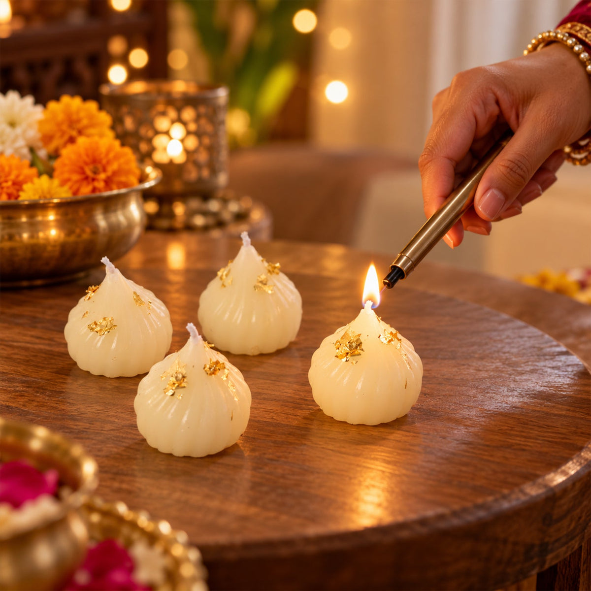 A hand lighting one of four white modak-shaped candles with gold flakes on a wooden table, surrounded by festive decor.