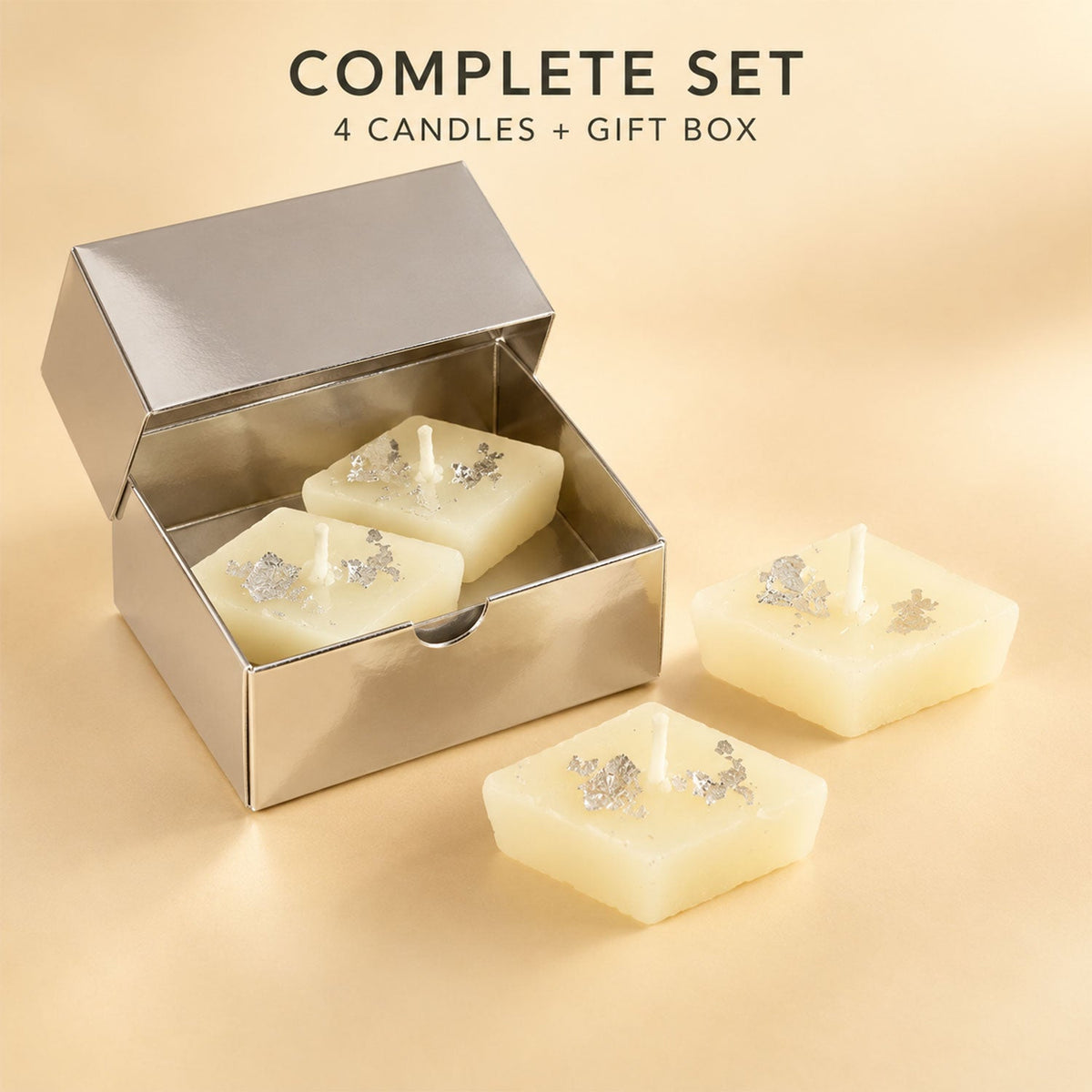 Two diamond-shaped cream candles with silver foil inside an open silver gift box, with two more candles placed outside the