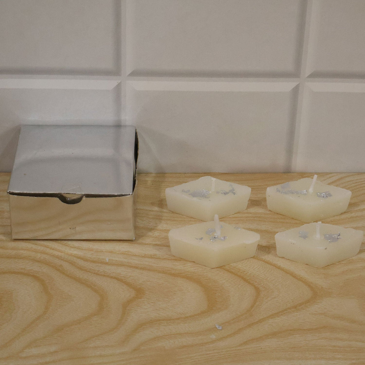 A silver gift box sits beside four diamond-shaped cream candles with silver foil on a light wooden surface, against a white