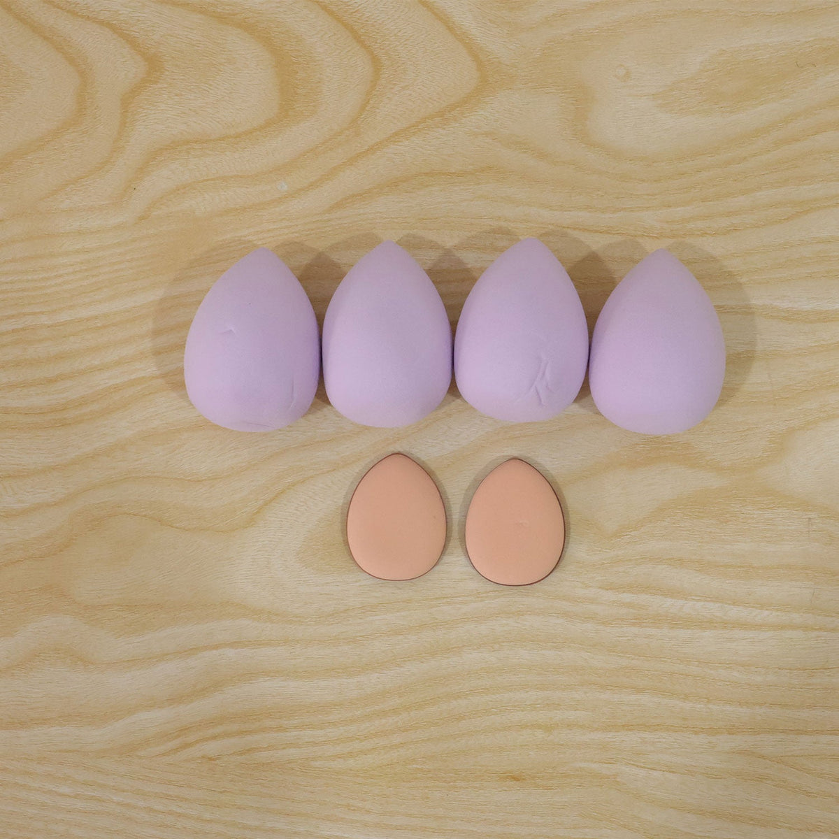 Four lavender teardrop sponges and two peach oval puffs are arranged on a light wooden surface.