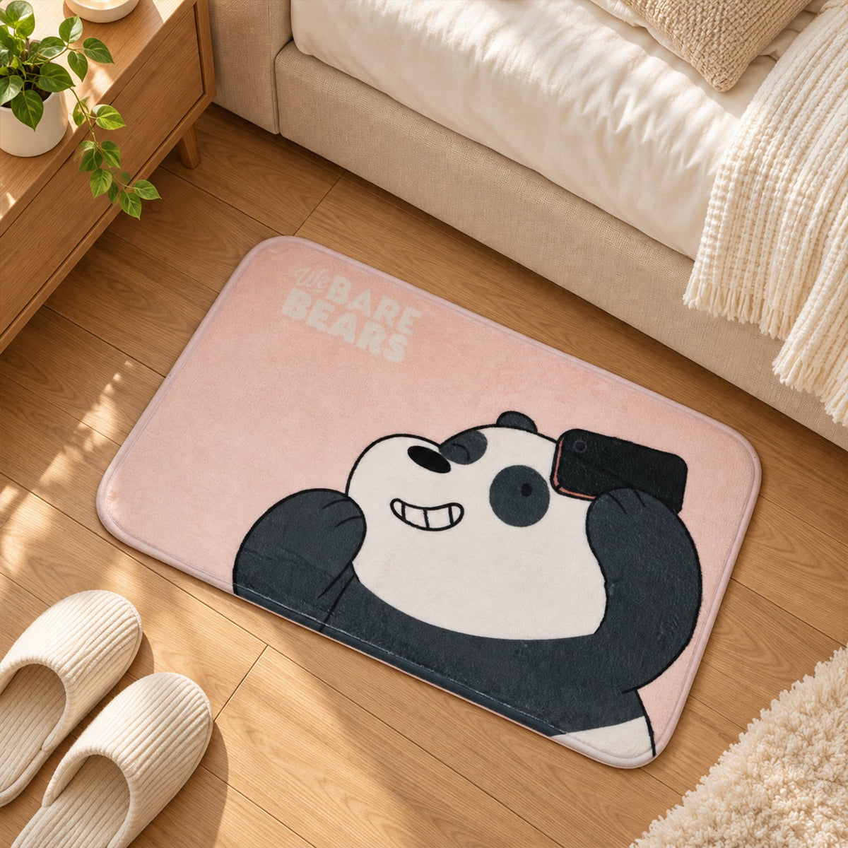 A pink panda mat lies on a wooden floor next to a bed, with a pair of light-colored slippers nearby.