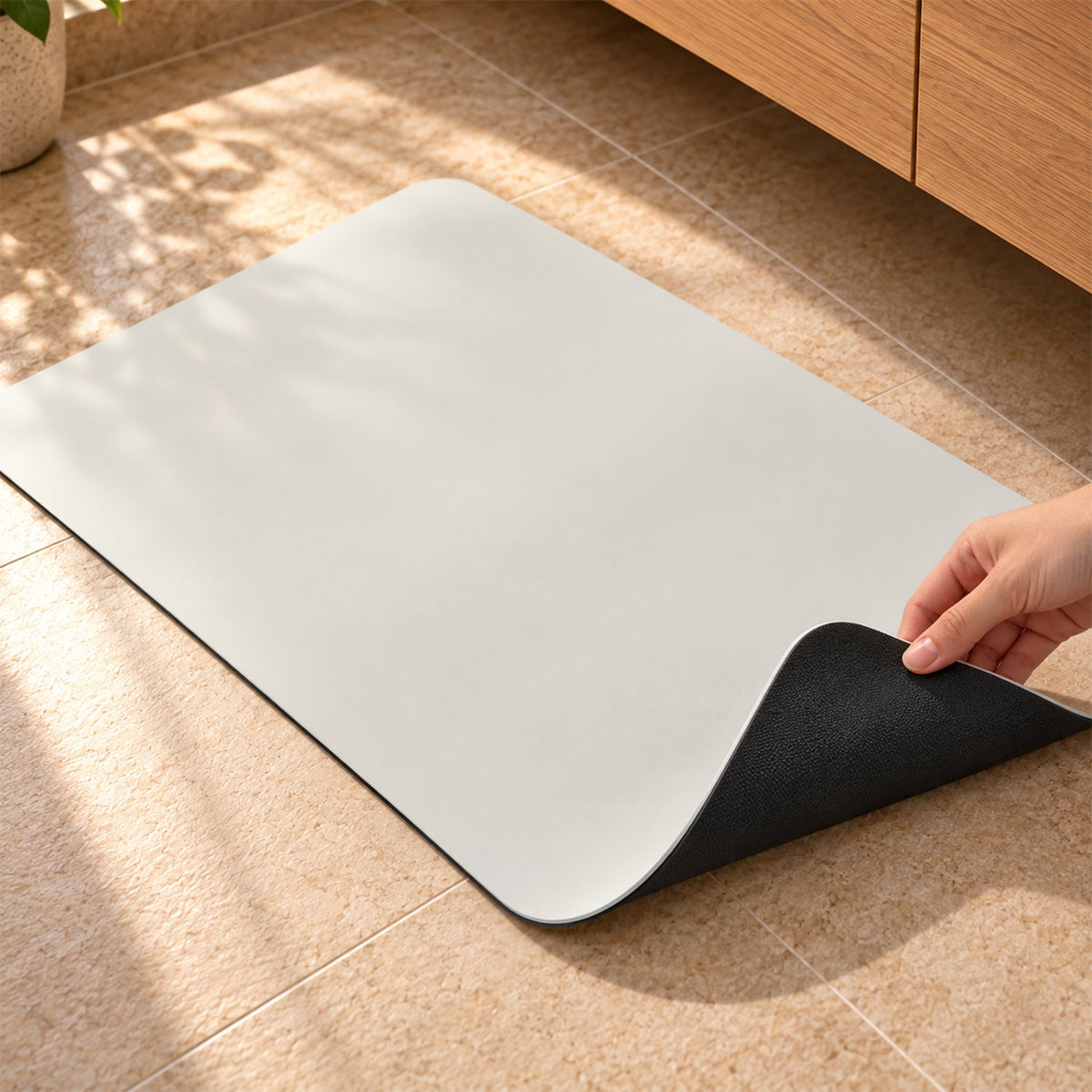 A hand lifts a corner of the cream mat, revealing its black non-slip underside on a tiled floor.