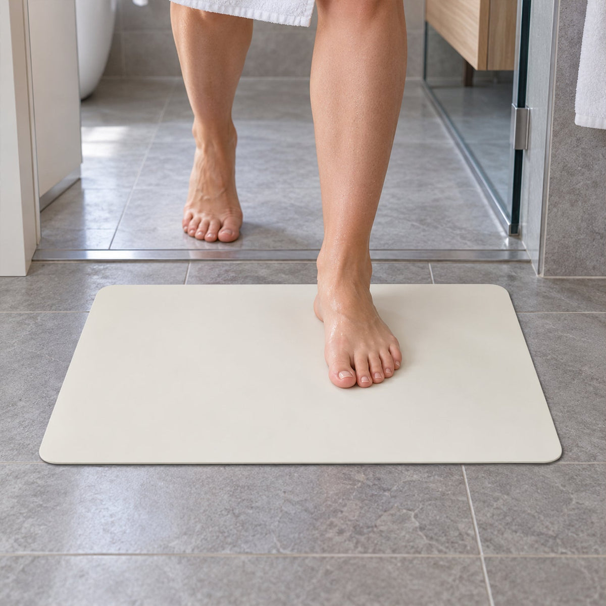 Bare feet step onto a cream rectangular mat on a grey tiled bathroom floor, near a glass shower enclosure.