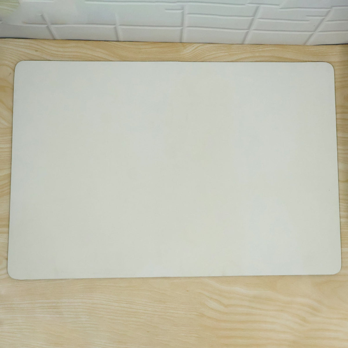 A cream rectangular mat with rounded corners lies flat on a light wood floor, against a white tiled wall.