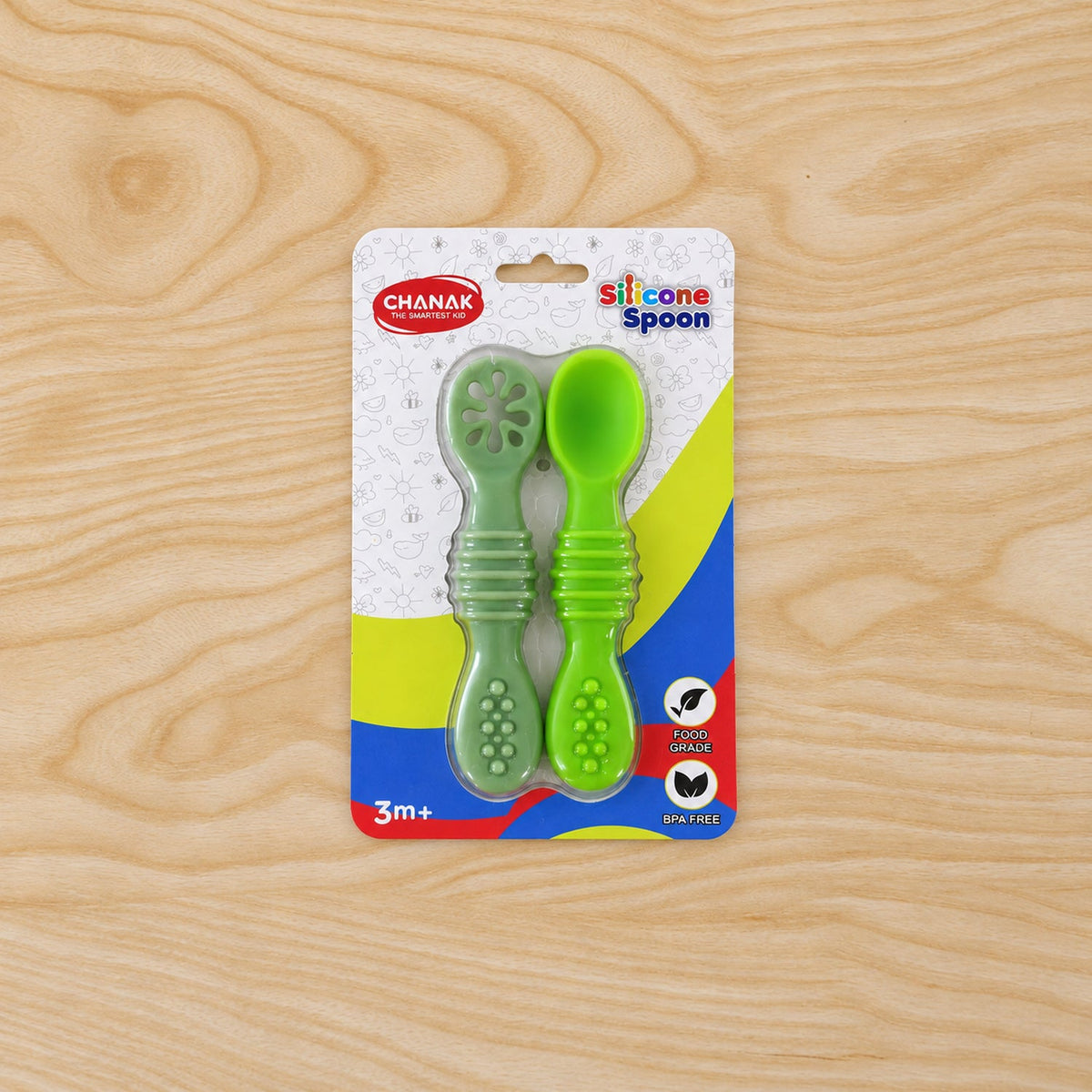 Two green silicone baby spoons in their clear plastic packaging, centered on a light wooden surface.