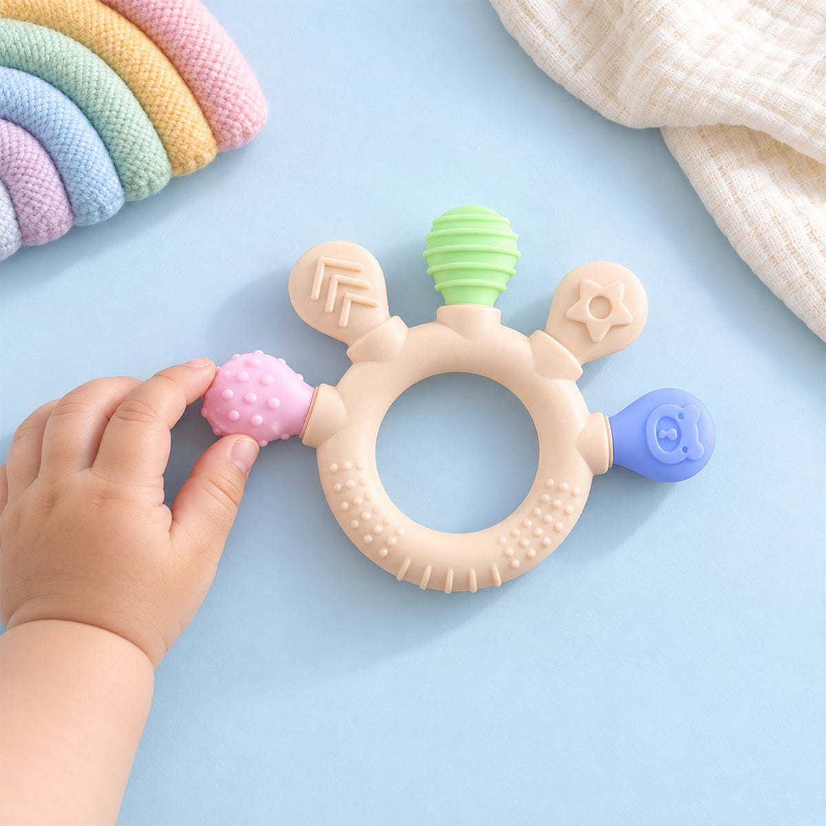 A baby's hand reaches for a beige teether with green, pink, and blue textured ends on a light blue surface.