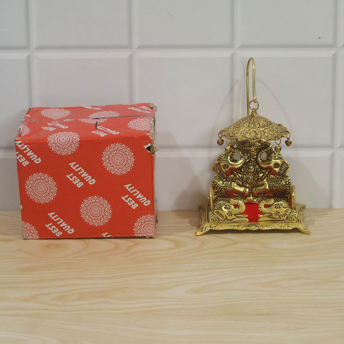 A gold-colored brass deity throne with an umbrella and red seat, placed next to its red and white patterned packaging box.