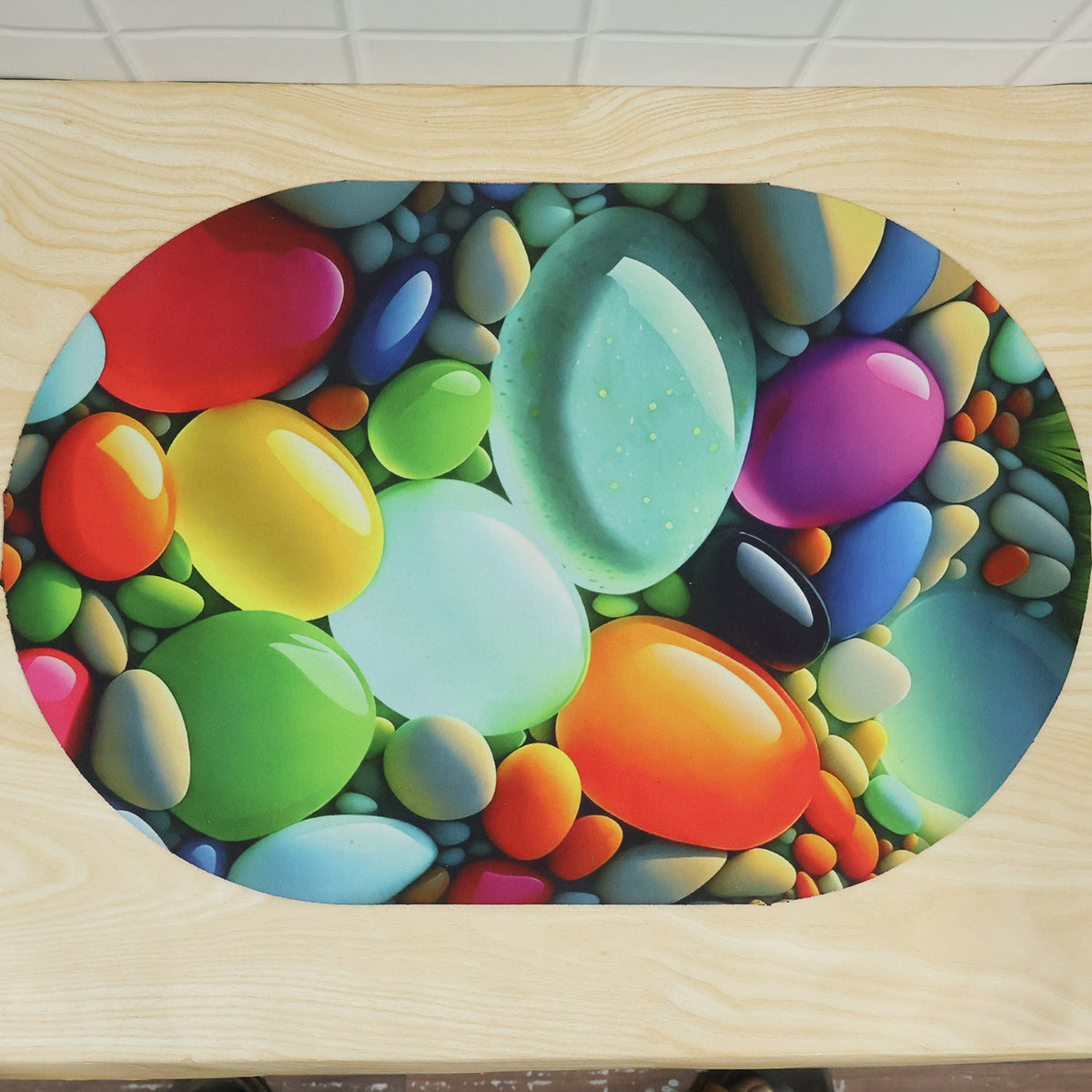 A top-down view of an oval mat with a bright, glossy pebble design on a light wooden surface.