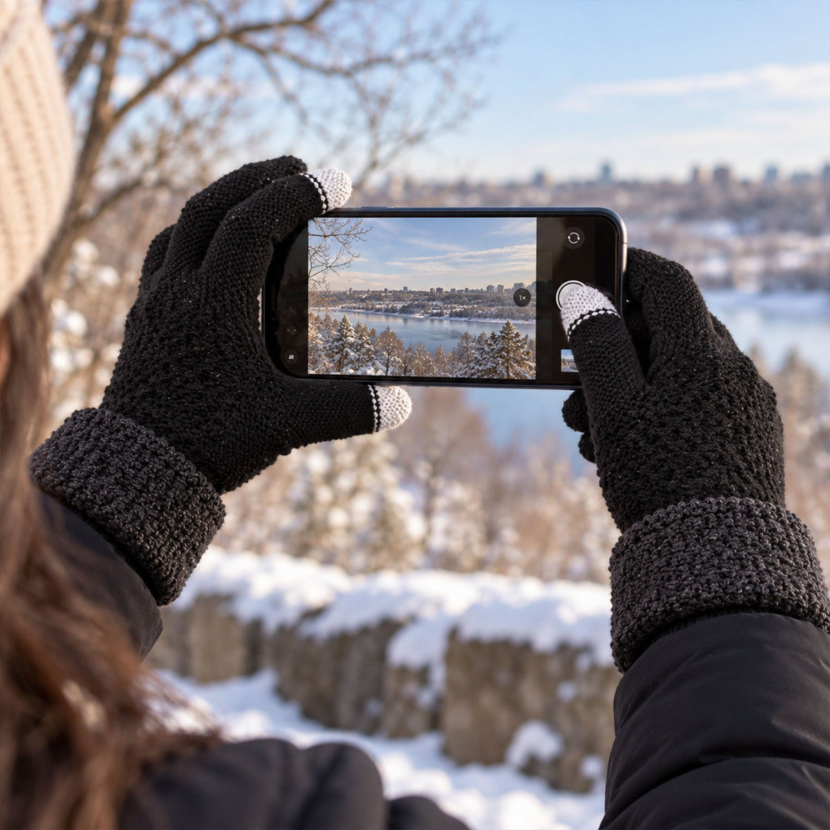Person wearing black knit gloves with gray cuffs and white touchscreen fingertips, holding a phone to capture a snowy
