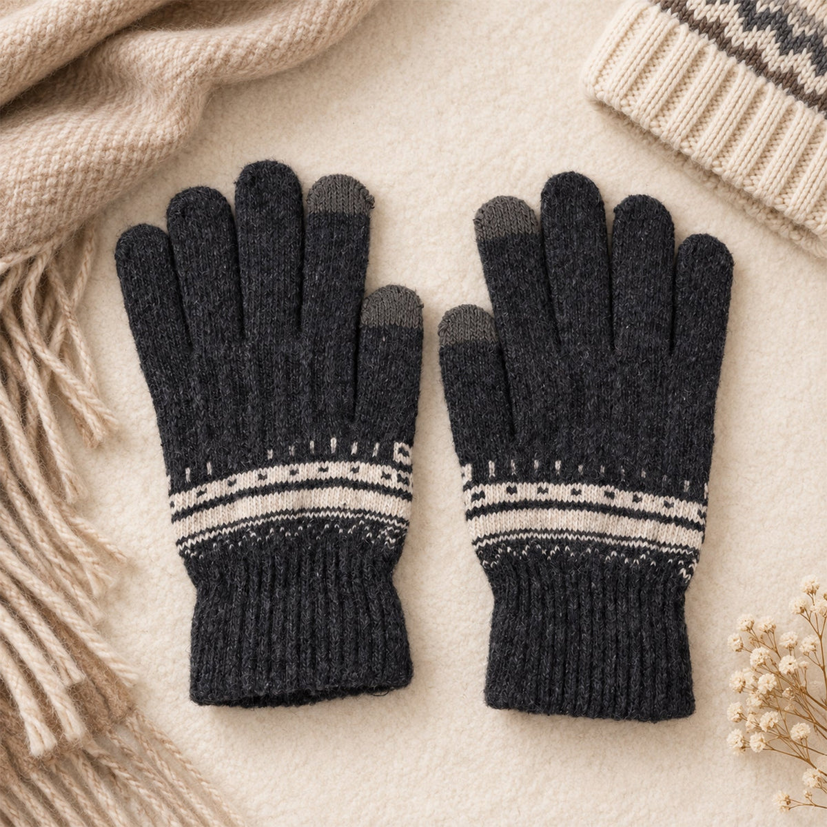 A pair of dark gray knitted gloves with white patterned cuffs lies flat on a light surface beside a beige scarf and hat.