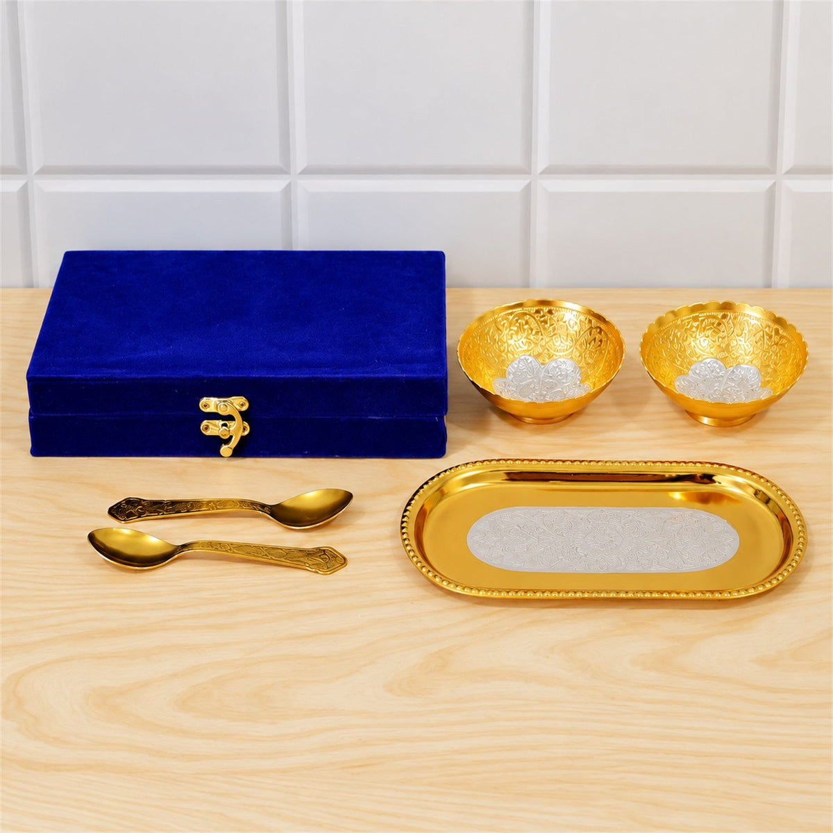 A blue velvet box, two golden bowls, two golden spoons, and a golden tray arranged on a light wooden surface.